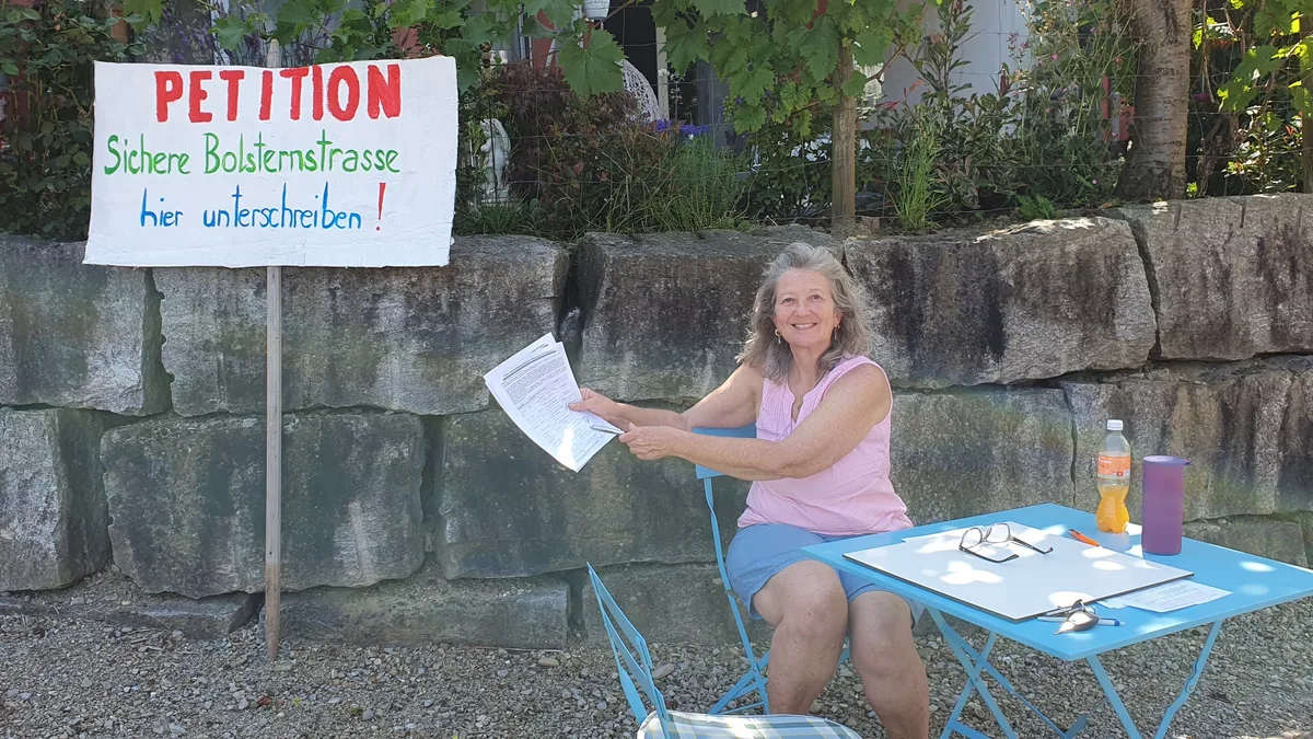 Man sieht eine Frau an einem Tisch mit einem Zettel in der Hand und einem Schild daneben.